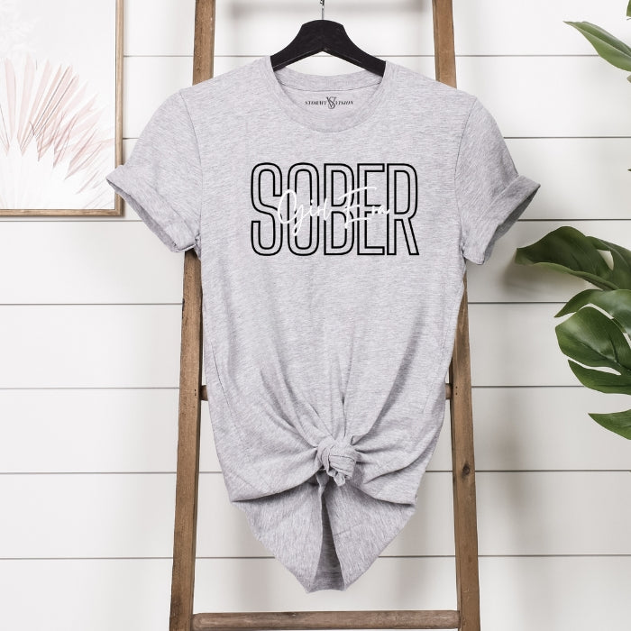Sober Girl Era Sports Grey Sobriety Tee For Women. Short Sleeve, Round Neck Tee with black and white font hanging from ladder. Front View.