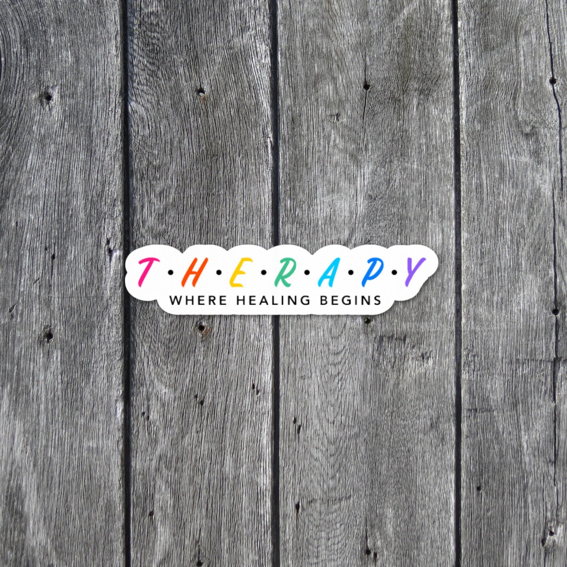Therapy Where Healing Begins - Inspirational Sticker is tiny and colorful. Placed on a grayish wooden background- close up.